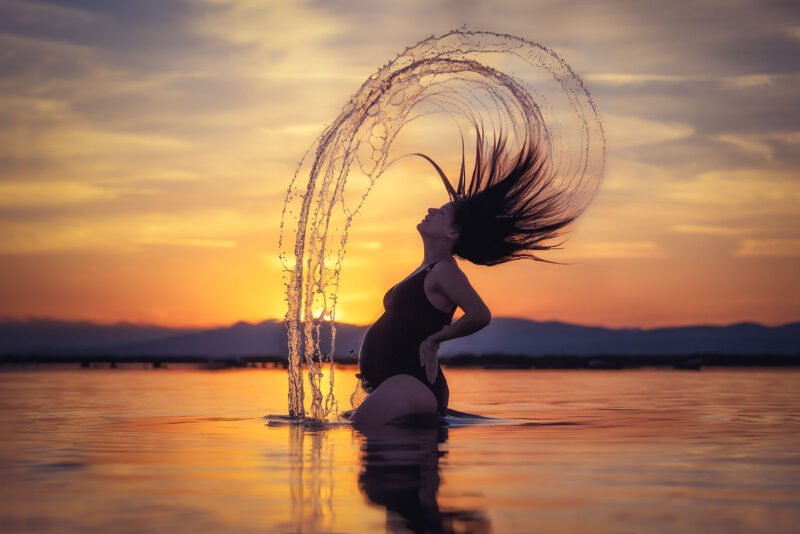 photo dune femme enceinte dans l'eau en contre jour au coucher de soleil avec les cheveux en mouvement créant une gerbe d'eau