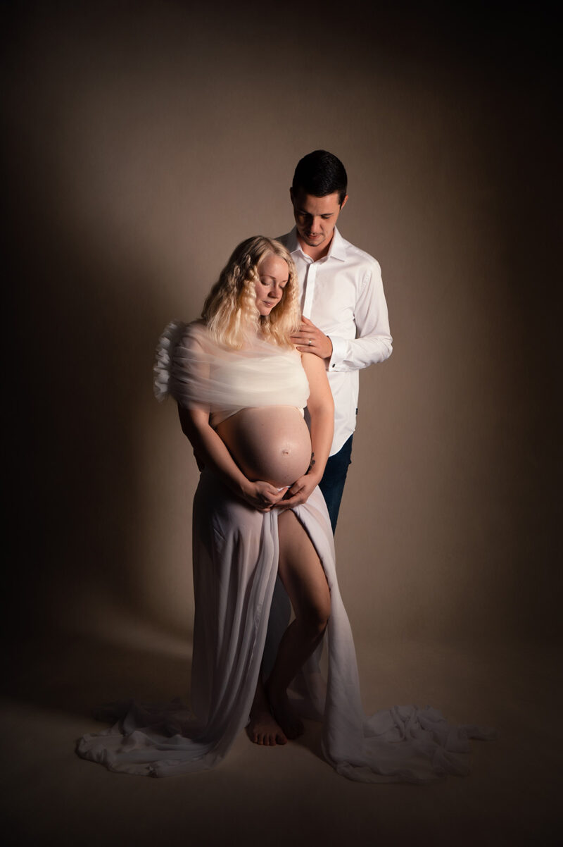 photo studio d'une femme enceinte habillée d'une robe blanche avec son conjoint posant la main sur son épaule
