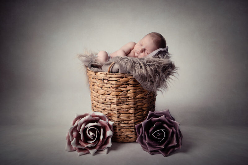 photo studio d'un bébé dans une corbeille dormant allongé sur le coté, deux grosses fleurs sont posés au sol