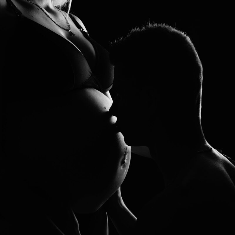 photo noir et blanc d'un homme embrassant le ventre de sa femme en clair obscur