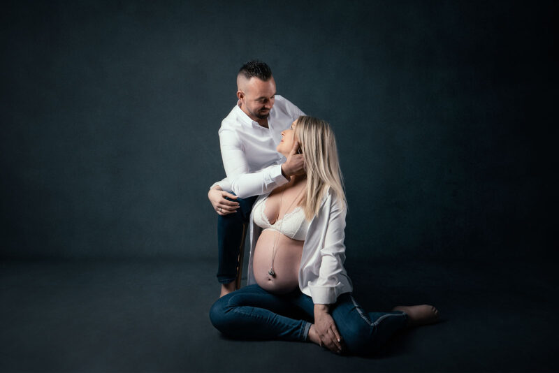 photo studio d'un couple dont la femme est enceinte. L'homme est assis sur une chaise et tiens le visage de la femme qui elle est assise au sol