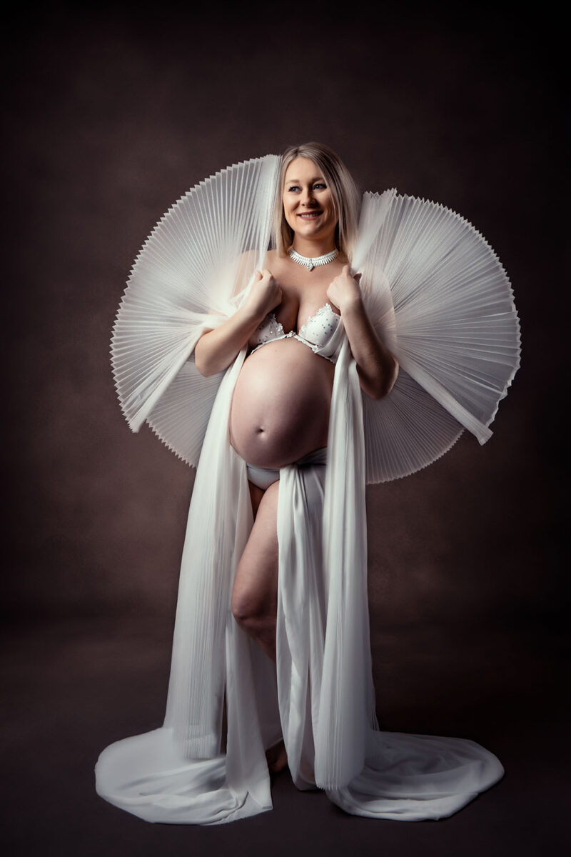 photo studio d'une femme blonde enceinte sur fond marron texturé vêtue d'une robe blanche