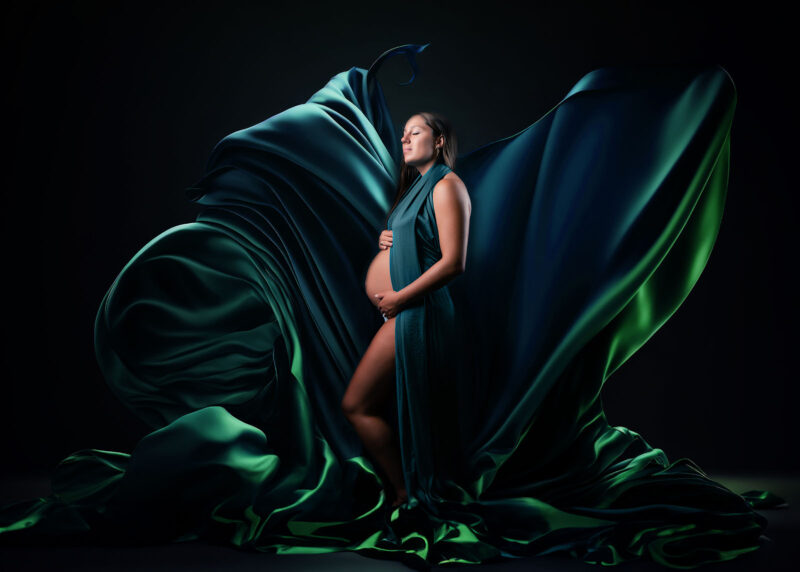 photo studio d'une femme enceinte aux cheveux longs vêtue d'un voile vert et bleu qui vole