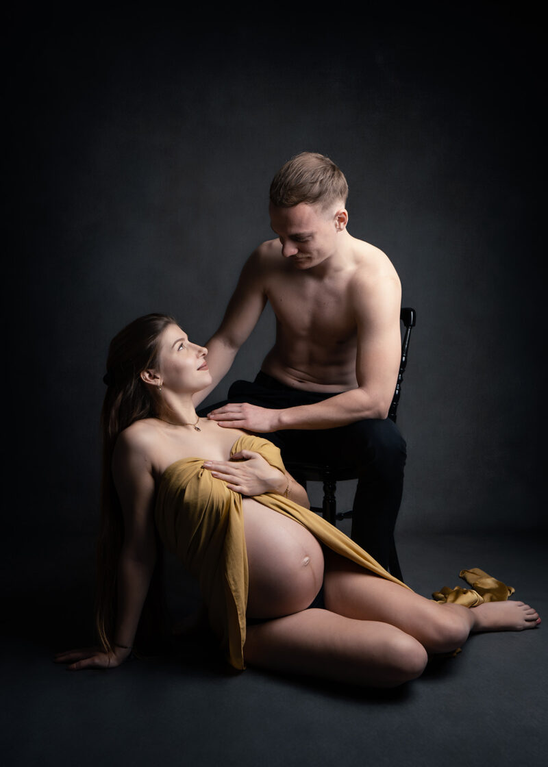 photo studio d'un couple où l'homme est assis sur une chaise et la femme au sol est enceinte