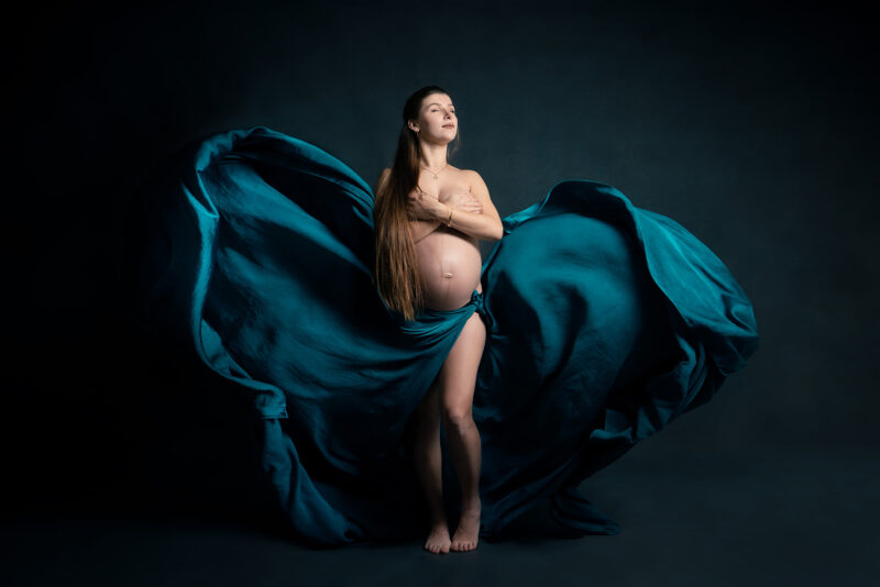 photo studio d'une femme enceinte portant un voile bleu volant au niveau de la taille et se couvrant la poitrine avec ses mains