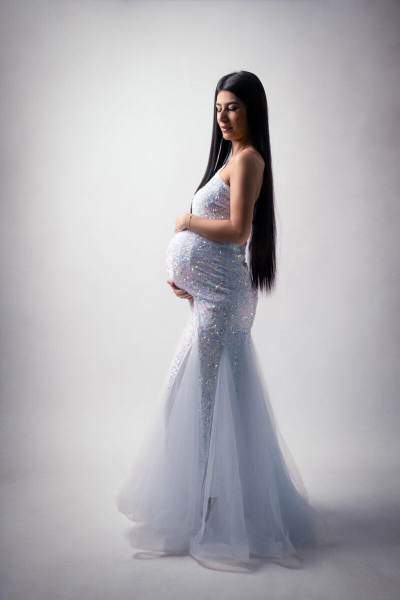 photo studio d'une femme enceinte de profil en plain pied portant une robe de soirée blanche pailletée