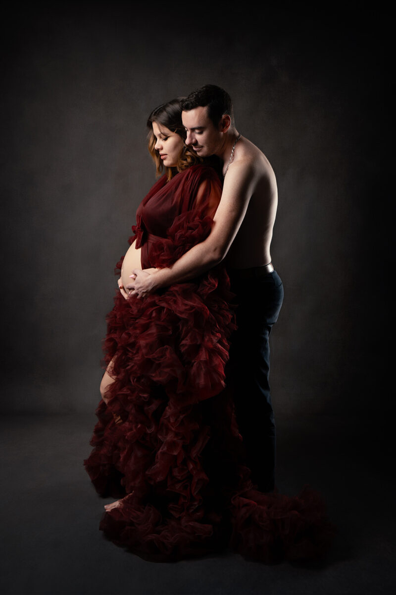photo studio de profil d'un couple dont la femme est enceinte et est vêtue d'une robe en tulle rouge. L'homme se tient derrière elle