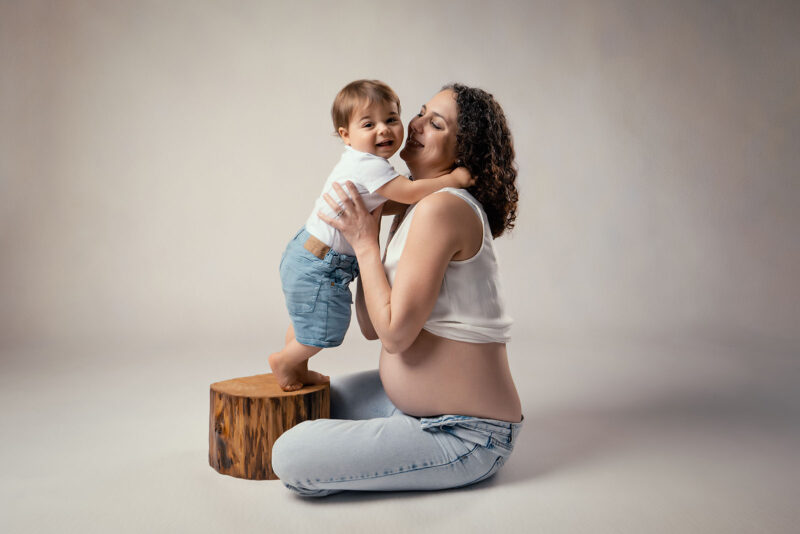 photo studio sur fond clair d'une femme enceinte assise et tenant son enfant dans les bras, celui ci est debout sur un tronc d'arbre