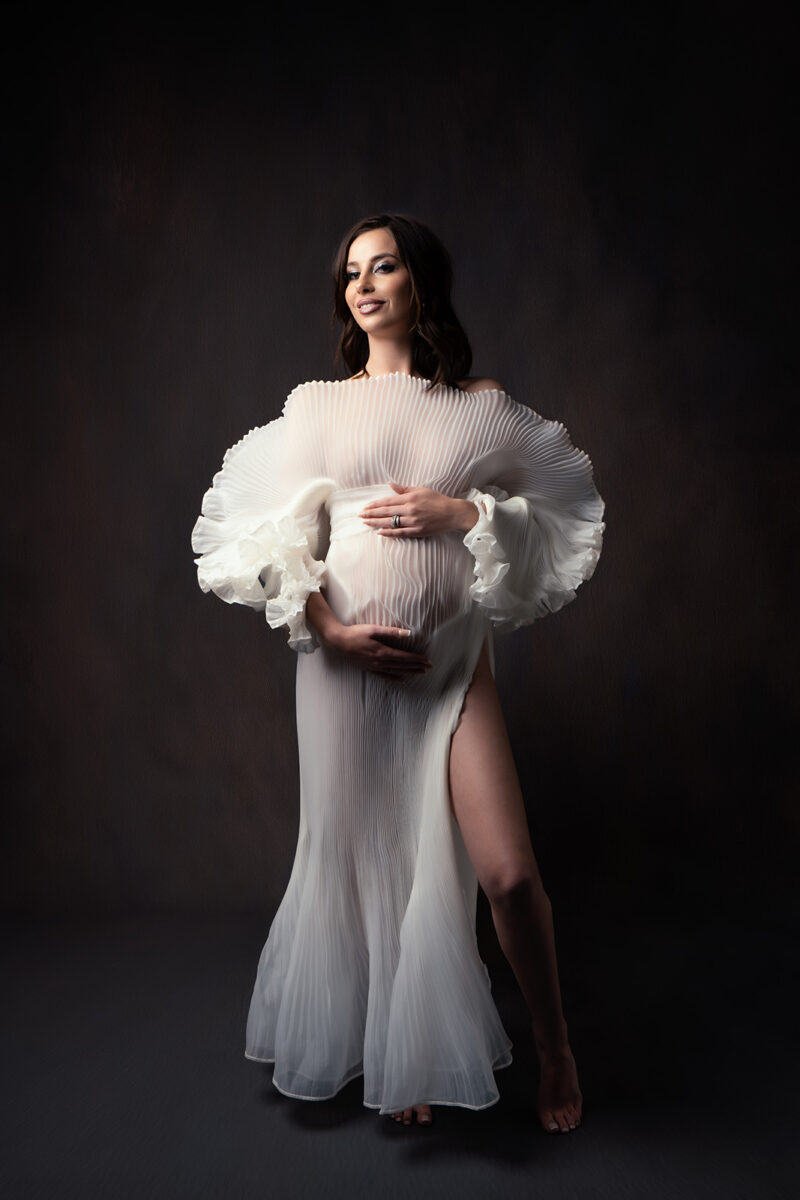 Photo studio d'une femme enceinte habillée d'une robe blanche en tulle avec une ouverture au niveau de la jambe