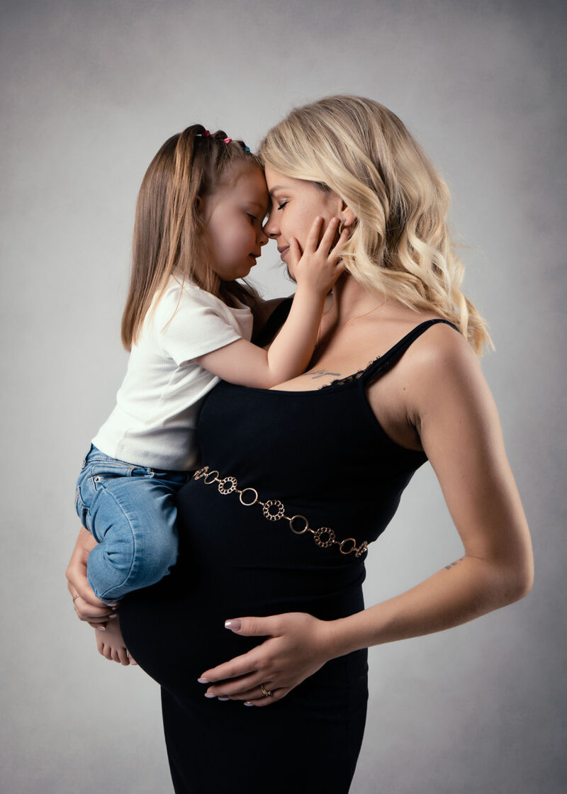 photo studio d'une femme enceinte blonde habillée d'une robe noire et tenant sa petite fille dans les bras