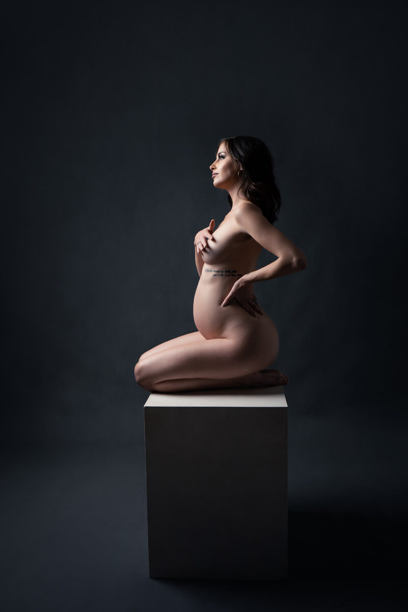 photo studio d'une femme enceinte nue se cachant la poitrine avec une main et assise sur un cube en bois blanc