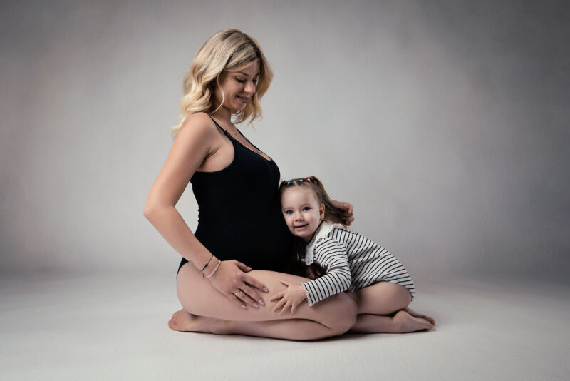 photo studio d'une femme enceinte assise avec sa petite fille posant sa tête sur son ventre