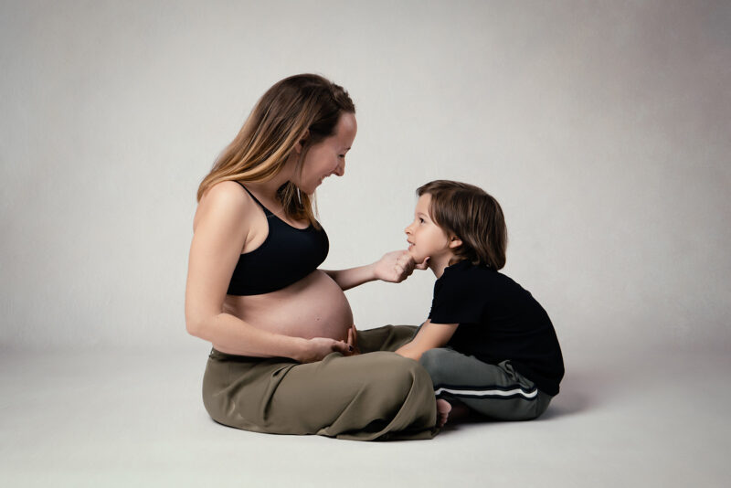 photo studio sur fond clair d'une femme enceinte assise et tenant le menton de son fils assis en face d'elle