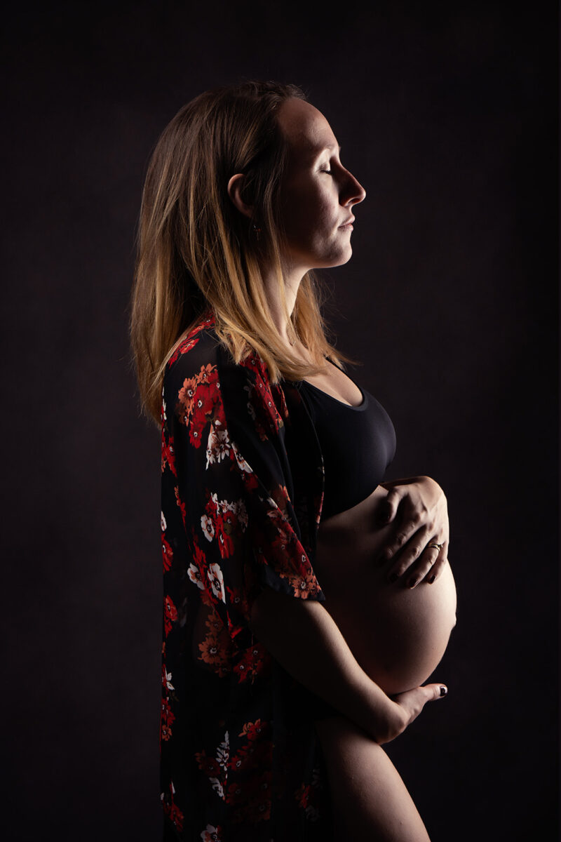 photo studio d'une femme enceinte en clair obscur, elle a les mains posé sur son ventre et ferme les yeux