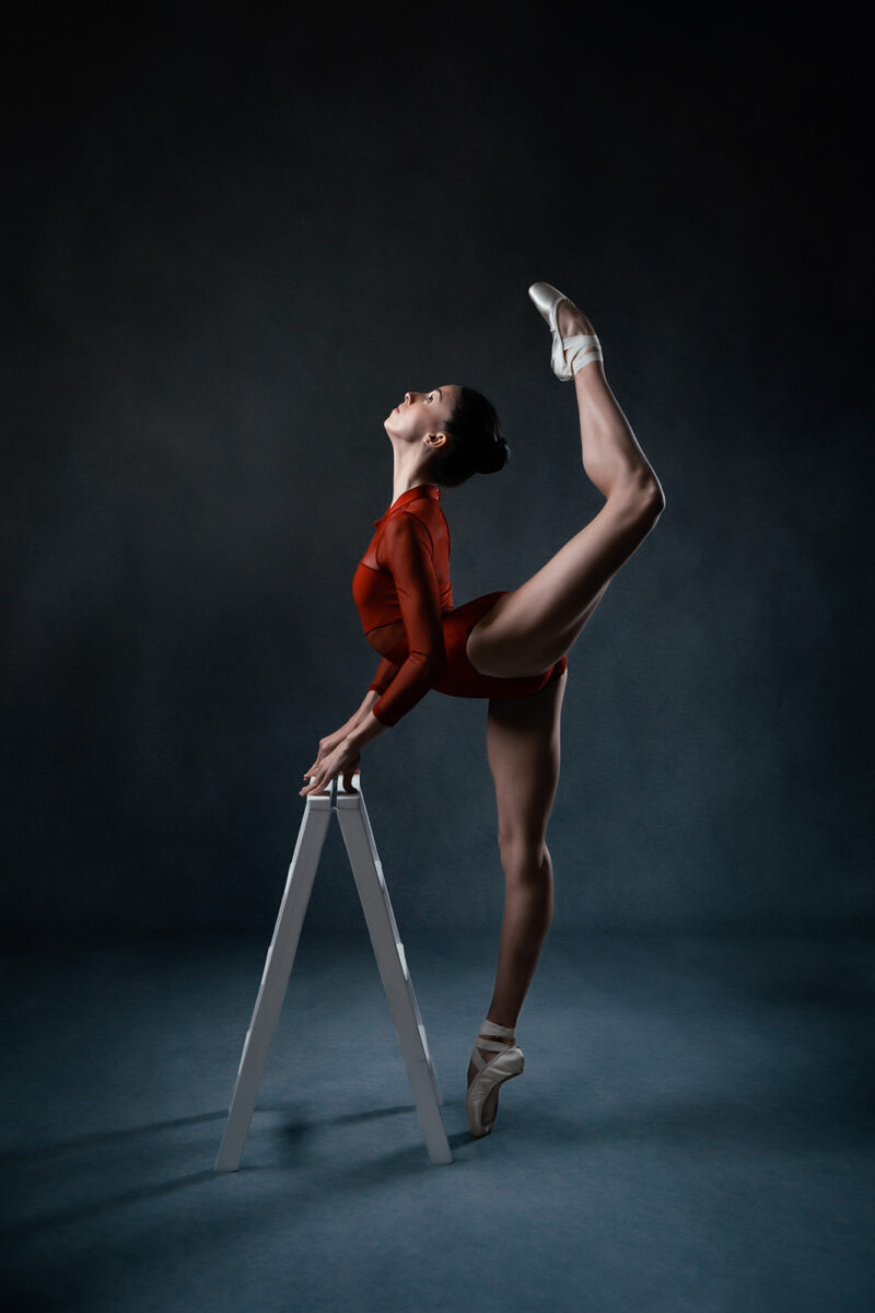 photo studio d'une danseuse classique habillée d'un body rouge et faisant une arabesque appuyée sur une petit échelle en bois