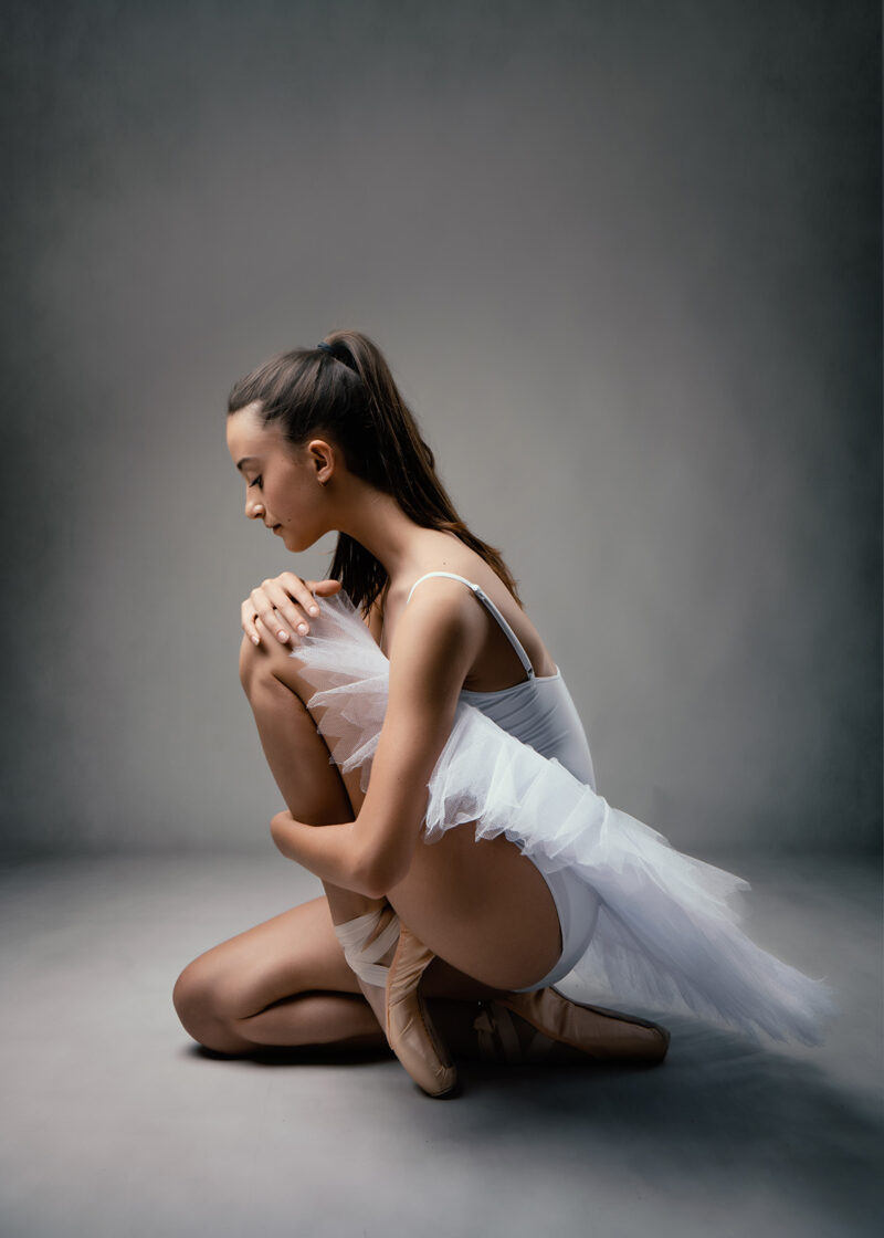 photo studio d'une danseuse classique habillée en tutu blanc assise au sol et tenant son genou