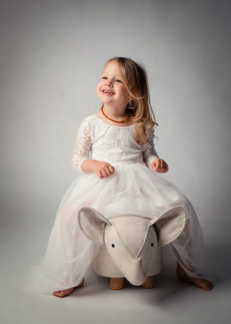 photo d'une petite fille habillée en robe blanche assise sur un petit tabouret en forme d'éléphant