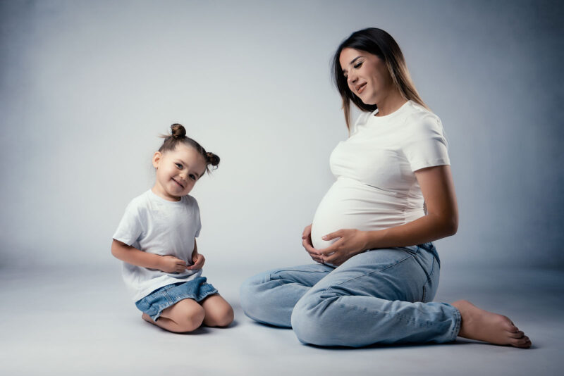 photo studio d'une femme enceinte assise au sol et tenant son ventre accompagnée de sa petit fille qui l'imite