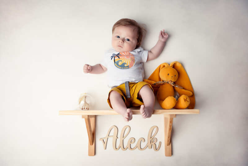 photo studio d'un bébé assis sur petite étagère en bois avec un doudou jaune posé à coté