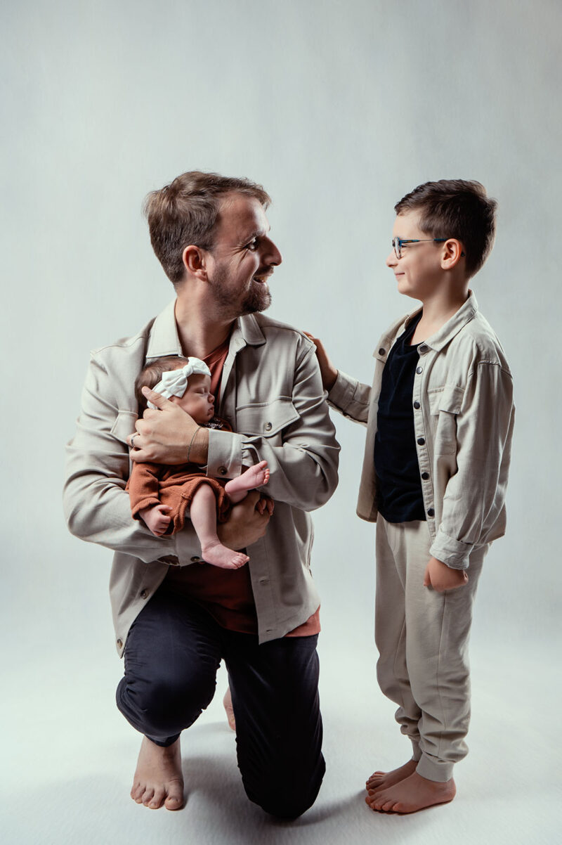 photo studio sur fond blanc d'un jeune papa tenant sa fille dans les bras avec un petit garçon se tenant debout à coté d'eux