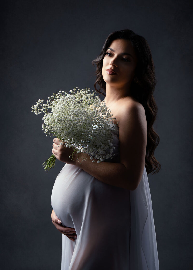 photo studio d'une femme enceinte vêtue d'un voile blanc et portant un bouquet de fleur à la main