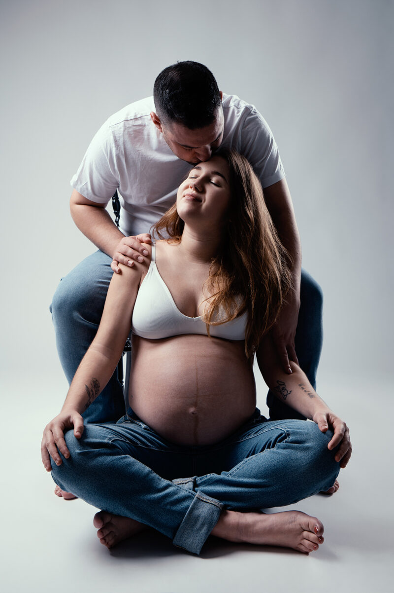 photo studio d'un couple dont la femme est enceinte et l'homme assis sur une chaise et lui embrasse le front