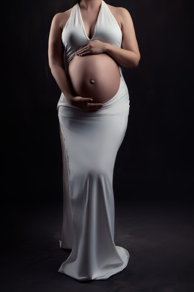 photo studio d'une robe blanche moulante d'une femme enceinte qui laisse découvrir le ventre mais dont on ne voit pas la visage