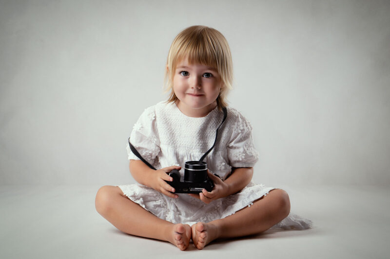 photo studio d'une petite fille blonde sur fond clair tenant un appareil photo dans ses mains