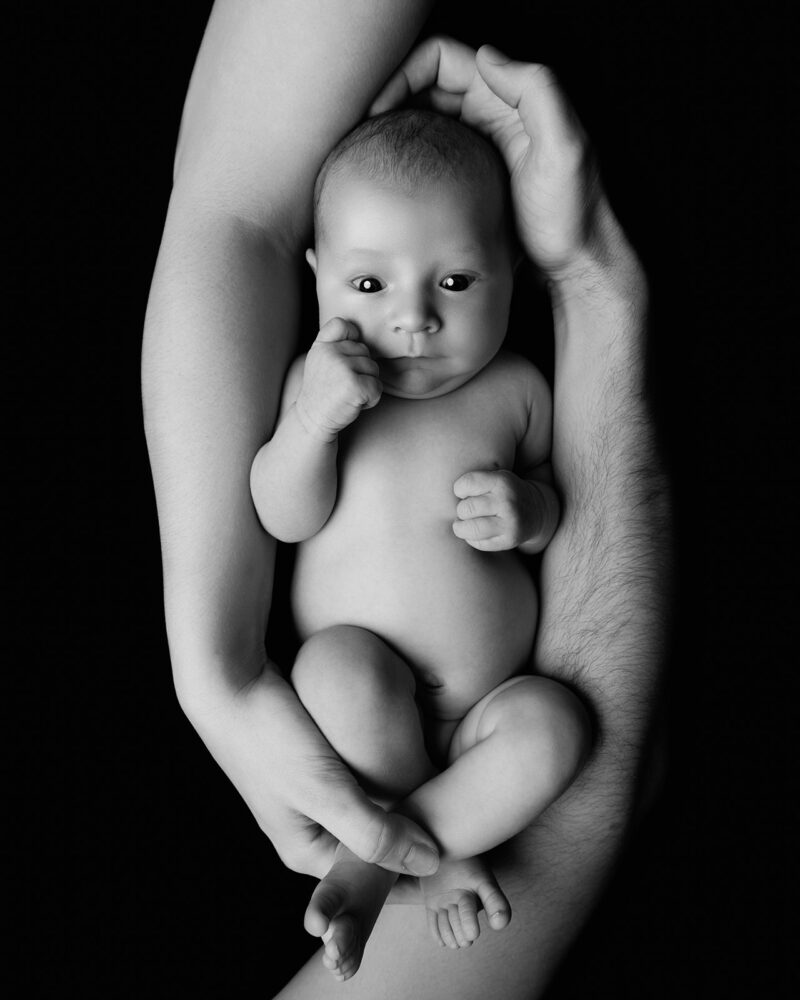 photo noir et blanc d'un bébé nu entouré des bras de ses parents