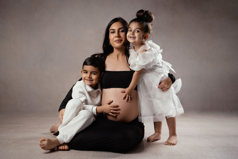 photo studio d'une femme enceinte assise au sol avec ses deux enfants dans les bras