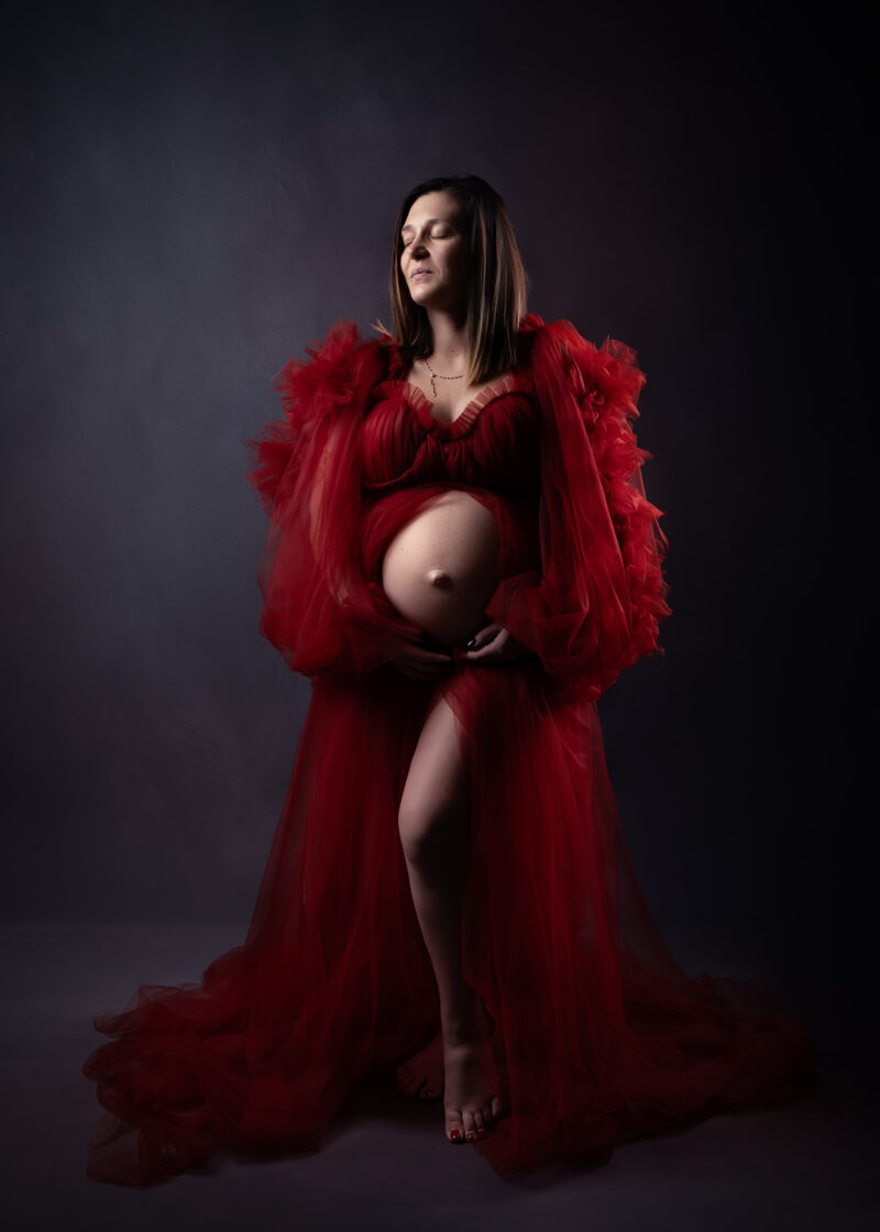 photo studio d'une femme enceinte sur fond foncé portant une robe en tulle de couleur rouge